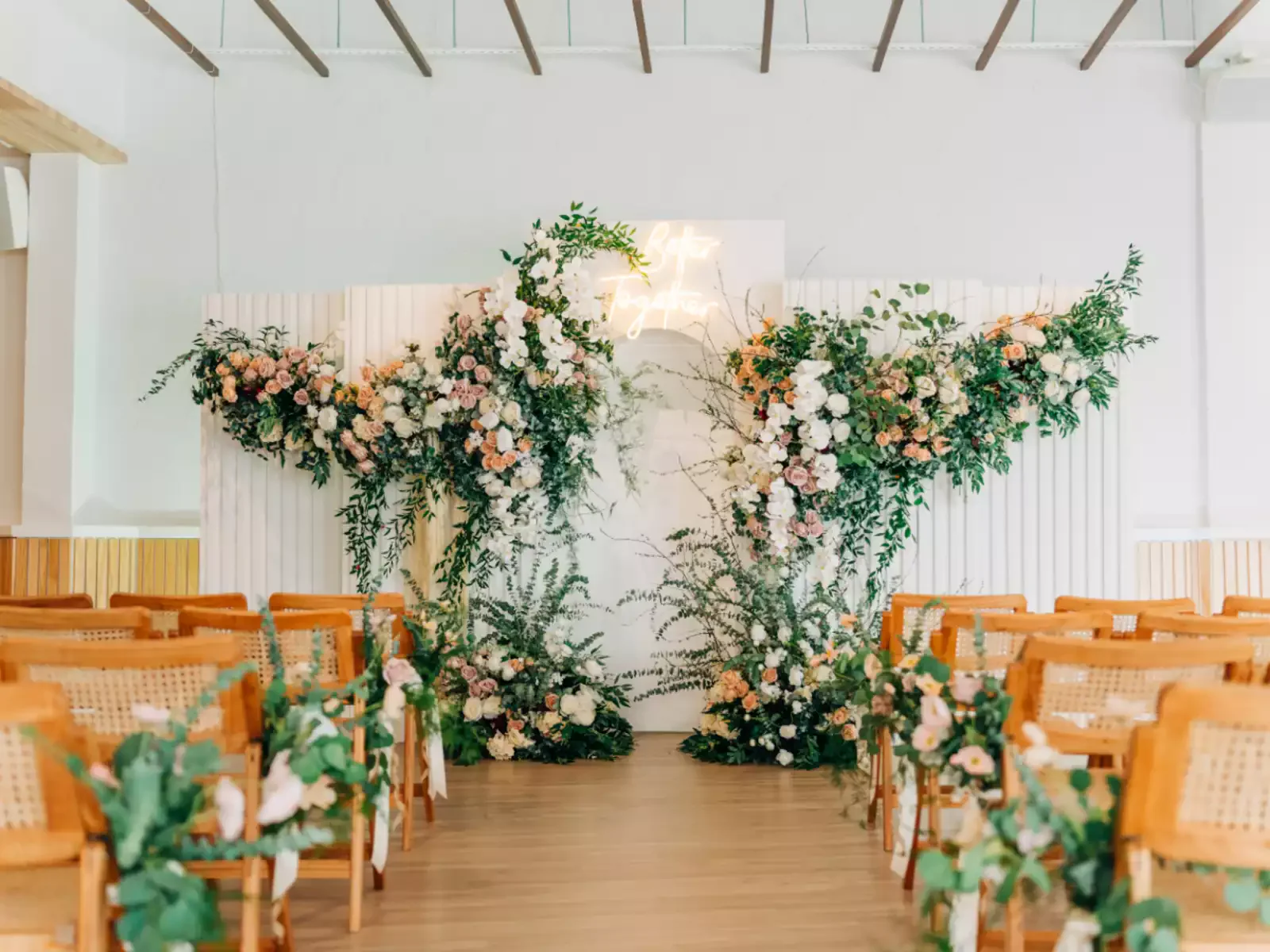Bright ceremony setup at Haus217 with a lush floral wall installation in peach, pink, white, and green, rattan chairs lining the aisle with floral accents